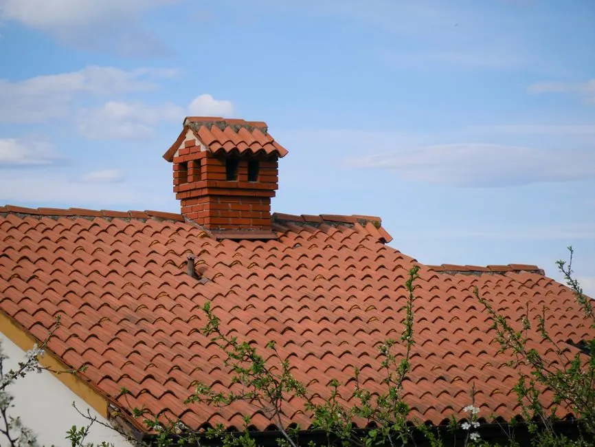 A charming red brick chimney stands atop a detailed clay-tiled roof under a clear blue sky.